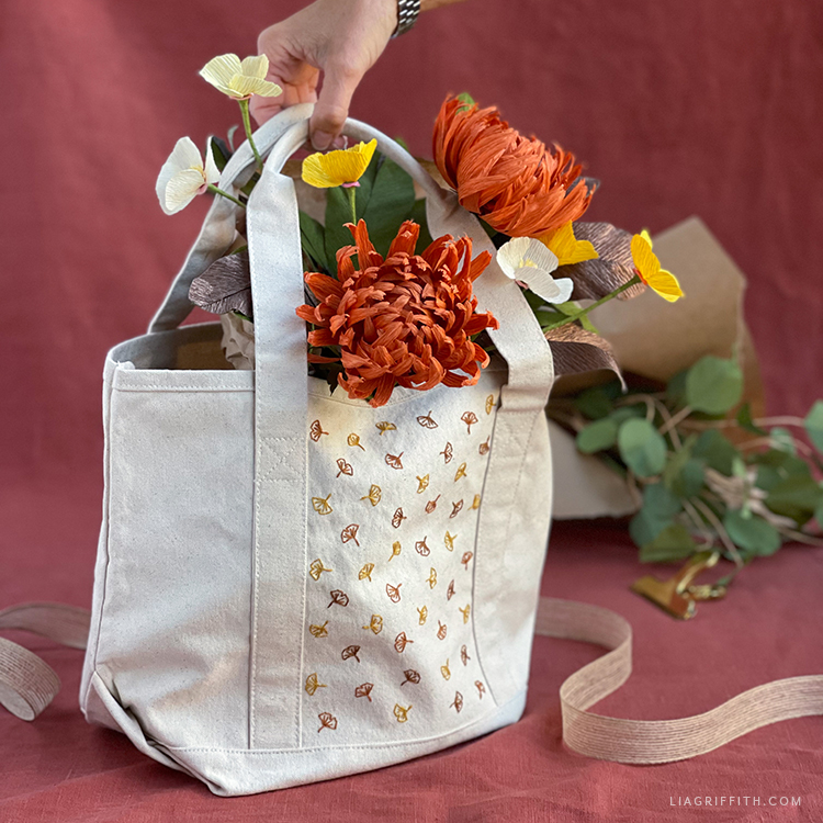 A large, white, patterned, canvas bag with a large assortment of paper and felt flowers. In the background is a paper bag of paper leaves and stems.
