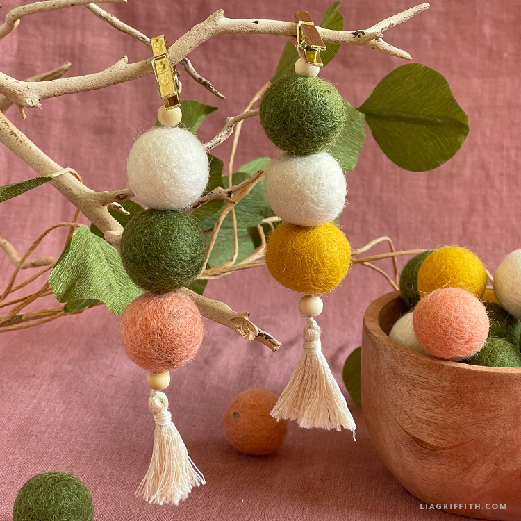 Two strings of three large felt balls hanging off of a tree branch next to a small wooden bowl of felt balls.